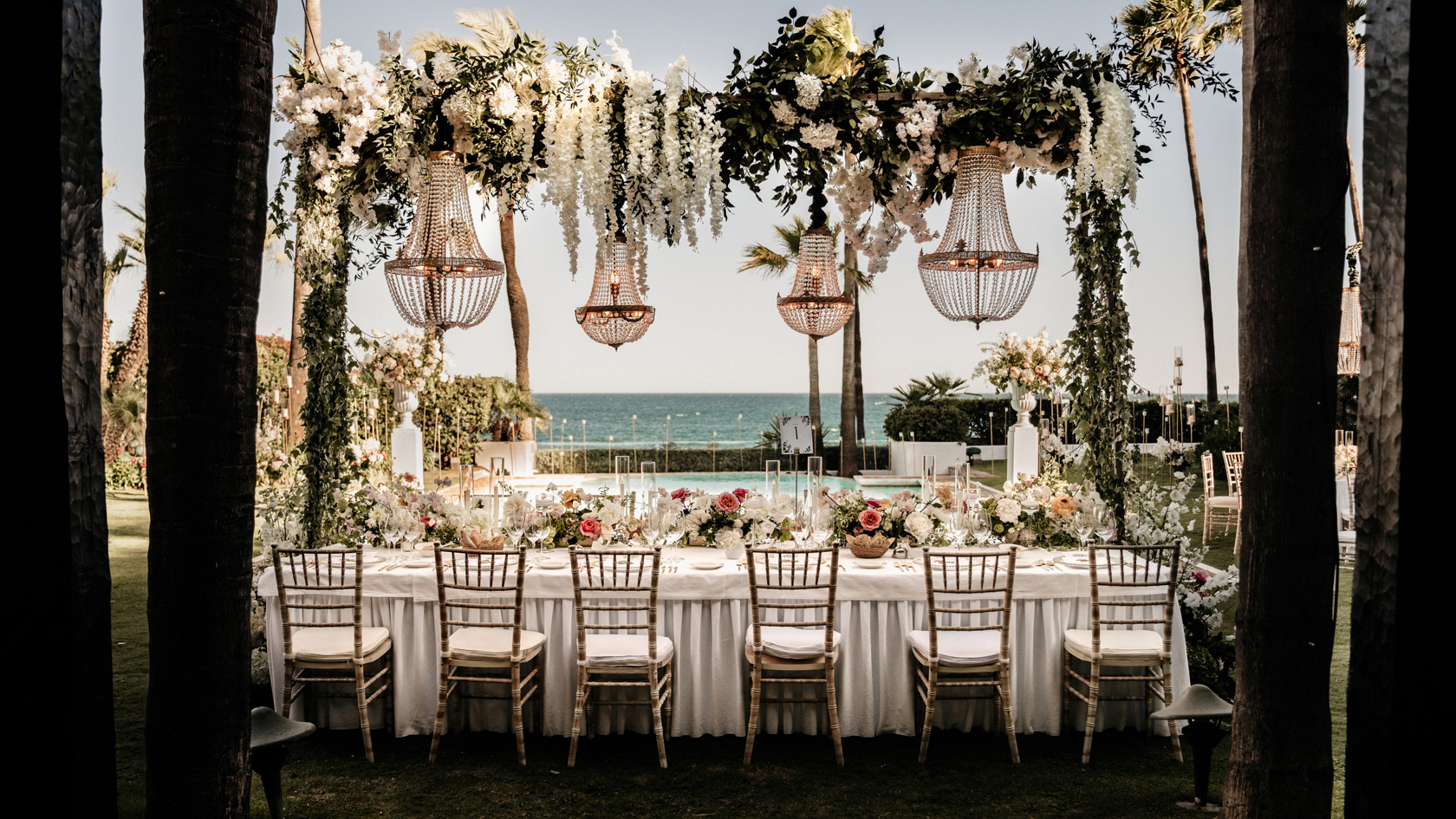 Een luxe bruiloftsdiner in Marbella, met een elegant gedekte lange eettafel onder een bloemenoverkapping met kristallen kroonluchters. De tafel is versierd met diverse bloemen en gedekt met fijn porselein, met uitzicht op een prachtig zeezicht en palmbomen op de achtergrond.